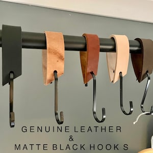 May include: A collection of genuine leather and matte black hooks hanging from a black metal rod. The leather hooks come in various colors, including black, tan, brown, and beige. The text "GENUINE LEATHER & MATTE BLACK HOOKS" is visible.