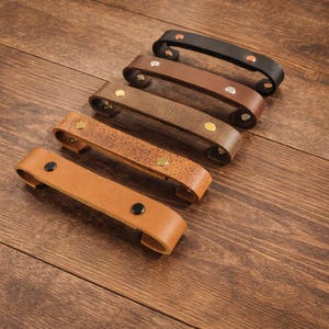 Könnte beinhalten: Eine Auswahl an Leder-Schubladengriffen in verschiedenen Braun- und Schwarztönen. Jeder Griff hat einen gebogenen Griff und ist mit Metallnieten befestigt. Die Griffe sind auf einer Holzoberfläche angeordnet, um ihre verschiedenen Farben und Texturen hervorzuheben.