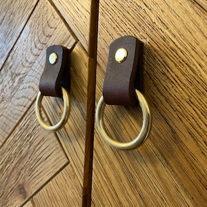 May include: Two dark brown leather cabinet pulls with brass ring handles and gold rivets. They are mounted on a wooden cabinet with a herringbone pattern. The cabinet is made of wood.