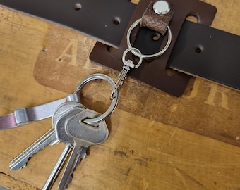 May include: A brown leather belt loop key holder with a silver key ring and a set of keys. The key holder is attached to a dark brown leather belt. The background is a wooden surface.