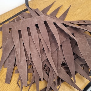May include: A pile of brown leather cutouts, each with a series of pointed edges and rectangular cutouts. The leather pieces are arranged on a wooden surface, creating a textured and layered effect. The cutouts appear to be for a craft project.