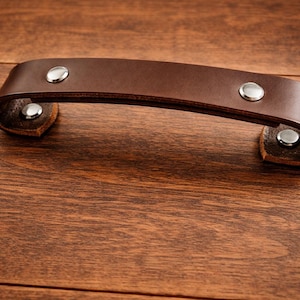 K&ouml;nnte beinhalten: Ein brauner Leder-Schrankgriff mit silberfarbenen Nieten. Der gebogene Griff ist an kleinen Lederlaschen befestigt. Die Hardware befindet sich auf einer dunklen Holzoberfl&auml;che.