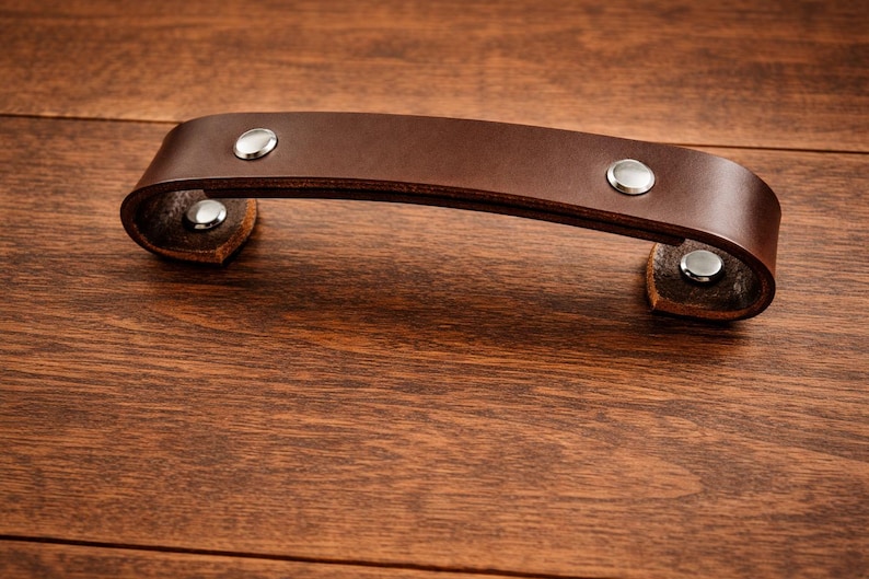 K&ouml;nnte beinhalten: Ein brauner Leder-Schrankgriff mit silberfarbenen Nieten. Der gebogene Griff ist an kleinen Lederlaschen befestigt. Die Hardware befindet sich auf einer dunklen Holzoberfl&auml;che.