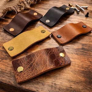 Może przedstawiać: Asortyment skórzanych uchwytów do szuflad w różnych kolorach: brązowym, czarnym, beżowym i złotym. Każdy uchwyt jest zakrzywiony i zabezpieczony metalowymi nitami. Uchwyty są ułożone na drewnianej powierzchni, prezentując ich fakturę i design.