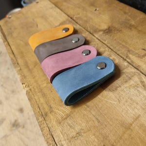 May include: Four leather key holders in shades of yellow, brown, pink, and blue. Each key holder is a curved rectangle with a metal snap closure. The key holders are arranged on a wooden surface.