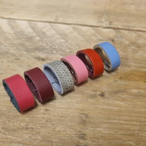 Puede incluir: Una colección de anillos de cuero coloridos en varios tonos, incluyendo rojo, burdeos, gris, rosa y azul. Los anillos están dispuestos sobre una superficie de madera, mostrando sus diferentes colores y texturas. Los anillos parecen hechos a mano.