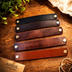 May include: Five rectangular leather wristbands in varying shades of black, dark brown, distressed reddish-brown, medium brown, and tan. Each band features two metallic gold-toned snap buttons at the ends for closure and a smooth leather texture.