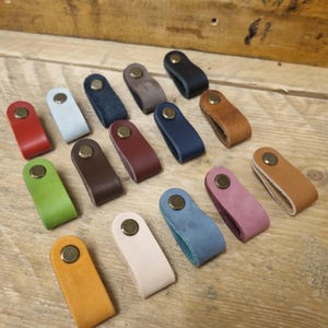 May include: A selection of leather drawer pulls in a variety of colours, such as red, blue, green, and brown. Each pull is secured with a brass-coloured rivet. The pulls are displayed on a wooden surface.