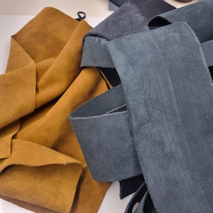 Puede incluir: Primer plano de piezas de cuero de ante marrón y gris. El cuero marrón está doblado, mientras que el cuero gris está cortado en tiras. Las texturas del cuero son visibles, destacando la suavidad y calidad del material. Ideal para manualidades.