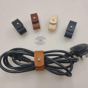 May include: A collection of leather cable organizers in black, brown, tan, and navy blue, each secured with a gold-colored snap. A brown leather organizer wraps around a black power cord. The Birmingham Leather Co. logo is visible.