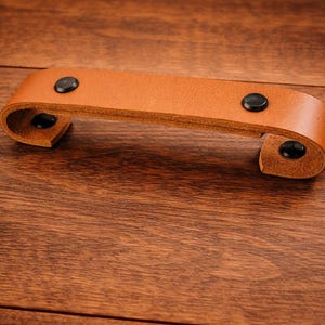 K&ouml;nnte beinhalten: Ein hellbrauner Leder-Schubladengriff mit schwarzen Nieten. Der Griff ist an beiden Enden gebogen und auf einer Holzoberfl&auml;che montiert. Die Hardware ist ein einfaches, aber elegantes Design.