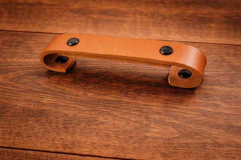 K&ouml;nnte beinhalten: Ein hellbrauner Leder-Schubladengriff mit schwarzen Nieten. Der Griff ist an beiden Enden gebogen und auf einer Holzoberfl&auml;che montiert. Die Hardware ist ein einfaches, aber elegantes Design.