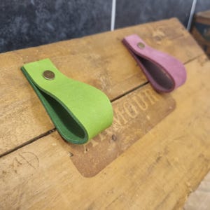 Op de afbeelding: Twee leren lade-grepen in limoengroen en mauve. Elke greep heeft een lusvorm, vastgezet met een koperkleurige klinknagel. De grepen liggen op een houten oppervlak, wat hun eenvoudige ontwerp en kleurcontrast laat zien.
