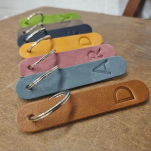 以下が含まれることがあります： 茶色、青、ピンク、黄色、緑など、さまざまな色の革製キーホルダーのコレクション。各キーホルダーには、シルバーのキーリングとエンボス加工された文字が付いています。キーホルダーは一列に並んでいます。