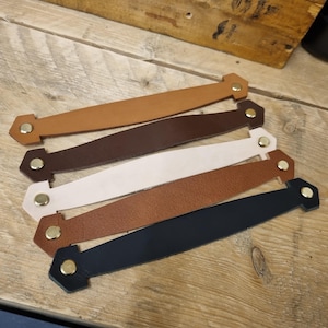 May include: Five leather drawer pulls in various colors: tan, dark brown, white, medium brown, and navy blue. Each pull has a rectangular shape with a hexagonal end and a gold-colored rivet.