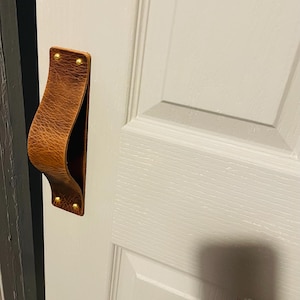 Może przedstawiać: Brązowa skórzana klamka do drzwi o zakrzywionym kształcie, przymocowana do białych drzwi za pomocą mosiężnych nitów. Drzwi są pomalowane na biało i mają panelowy design.
