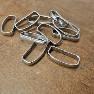 May include: A collection of silver metal belt loops, scattered on a brown leather surface. The loops are rectangular with rounded edges and a hinged design, suggesting they are designed for use with belts or straps.