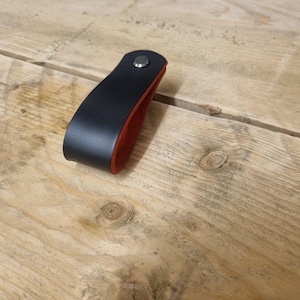May include: A black leather cabinet pull with a red interior, secured with a silver rivet. The pull is curved and sits on a wooden surface.