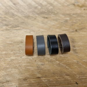 Puede incluir: Cuatro anillos de cuero en tonos marrón, gris, negro y marrón oscuro, dispuestos sobre una superficie de madera. Los anillos son de igual tamaño y parecen estar hechos de un material de cuero liso y duradero. Los anillos son probablemente para manualidades o joyería.