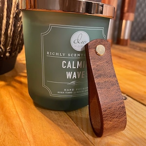 以下が含まれることがあります： 銅製の蓋と茶色の革製の取っ手が付いた緑色のガラス製のキャンドルジャー。キャンドルには「Richly Scented Calming Waves」と「Hand Poured」のラベルが貼られています。