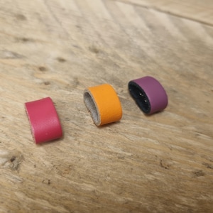 Puede incluir: Tres pequeños anillos de cuero en fucsia, naranja y morado. Los anillos están colocados sobre una superficie de madera, mostrando sus colores vibrantes y su diseño sencillo. El cuero parece liso y los bordes están cuidadosamente acabados. Cada anillo mide unos 2,5 cm de diámetro.