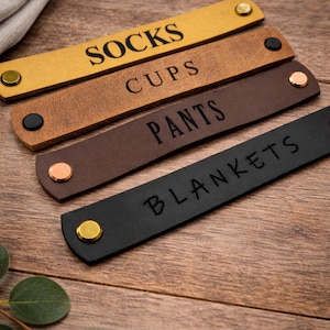 May include: Four leather labels in shades of gold, brown, and black, each with a different word: SOCKS, CUPS, PANTS, and BLANKETS. The labels have metal rivets and are arranged on a wooden surface.