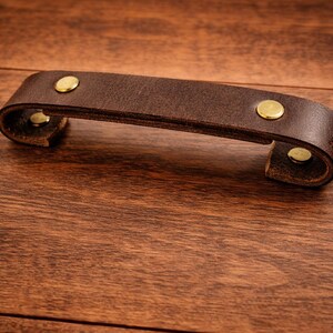 K&ouml;nnte beinhalten: Ein dunkelbrauner Leder-Schubladengriff mit Messingnieten. Der Griff ist gebogen und scheint aus dickem, hochwertigem Leder gefertigt zu sein. Die Beschl&auml;ge haben eine warme Goldfarbe, die mit dem dunkelbraunen Leder kontrastiert.
