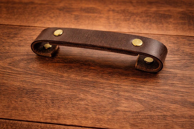K&ouml;nnte beinhalten: Ein dunkelbrauner Leder-Schubladengriff mit Messingnieten. Der Griff ist gebogen und scheint aus dickem, hochwertigem Leder gefertigt zu sein. Die Beschl&auml;ge haben eine warme Goldfarbe, die mit dem dunkelbraunen Leder kontrastiert.