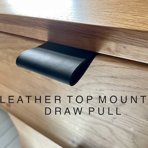 May include: A black leather top-mounted drawer pull is attached to a light-colored wooden drawer. The text "LEATHER TOP MOUNTED DRAW PULL" is displayed below the drawer pull.
