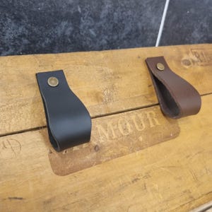 May include: Two leather drawer pulls, one black and one brown, each attached with a brass rivet. The pulls are on a wooden surface with the word "MOUR" visible. The pulls are designed for cabinets or drawers.