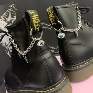 May include: Black leather boots with yellow stitching and a chain with an evil eye charm attached to the ankle. The boots have a black strap with the text "Lil Wait" on it.