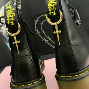 May include: Black leather boots with yellow stitching and a gold-colored sword charm hanging from the pull tab. The pull tab has the text "Air Wair" in yellow. The boots are on a pink surface.