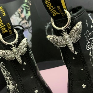 May include: Black leather boots with white embroidery of clouds and stars. The boots have silver charms in the shape of moths attached to the laces. The laces are black with yellow text that reads "with bouncing soles".