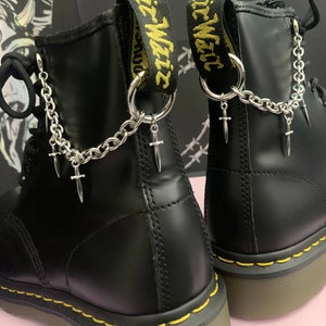 May include: Black leather boots with yellow stitching and a silver chain with small cross charms attached to the top of the boots. The boots have a yellow and black "Wait" strap on the back.