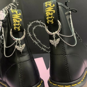 May include: A pair of black leather boots with yellow stitching and silver chains and charms attached to the laces. The charms are in the shape of a skull and wings. The boots have a black strap with the text "It. Waits" in yellow lettering.