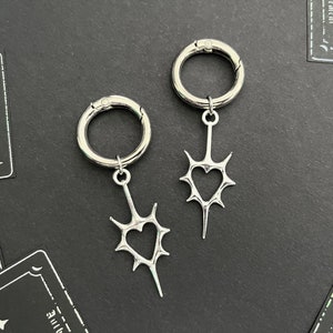 May include: Two silver hoop earrings with a heart-shaped design and a spiky, thorny detail. The earrings are on a black background.