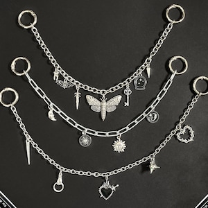 May include: Three silver chain necklaces with various charms, including a moth, key, moon, sun, and star. Each chain has a large ring at the top. The background is black with decorative cards.