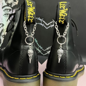 May include: Black leather boots with yellow stitching and a silver chain with a skull pendant attached to the back of each boot. The chain is attached to a yellow strap with the text "Air Waiz" on it.