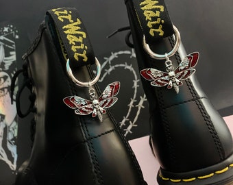 Puede incluir: Botas de cuero negro con costuras amarillas y un amuleto plateado sujeto a los cordones. El amuleto es una polilla roja y plateada con una calavera en su cuerpo. Las botas tienen una correa negra con el texto "It Wait" en amarillo.