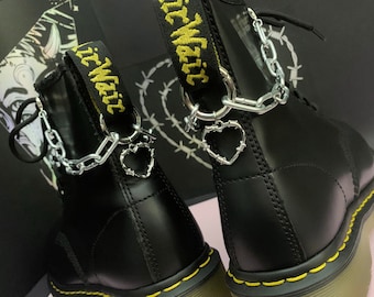 May include: Black leather boots with yellow stitching and a thick sole. The boots have a silver chain and heart charm attached to the laces. The boots have a yellow embroidered strap with the text "It Wait" on each boot.
