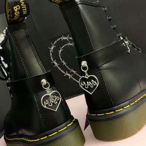 May include: A pair of black leather boots with yellow stitching and a black leather strap with a silver heart charm. The heart charm has a flame design.