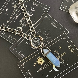 May include: A silver chain necklace with a crystal pendant. The pendant is a light blue, pointed crystal with a silver cap. The necklace is laying on top of several tarot cards. The cards are black with white writing and symbols. The writing on the cards says "Antho", "The Sun", "Temper", and "Judgement".