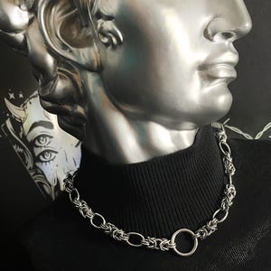 K&ouml;nnte beinhalten: Eine silberne Kettenkette mit einem gro&szlig;en, runden, silbernen Ringverschluss. Die Kette wird von einer Person getragen, die ein schwarzes Rollkragenhemd tr&auml;gt.