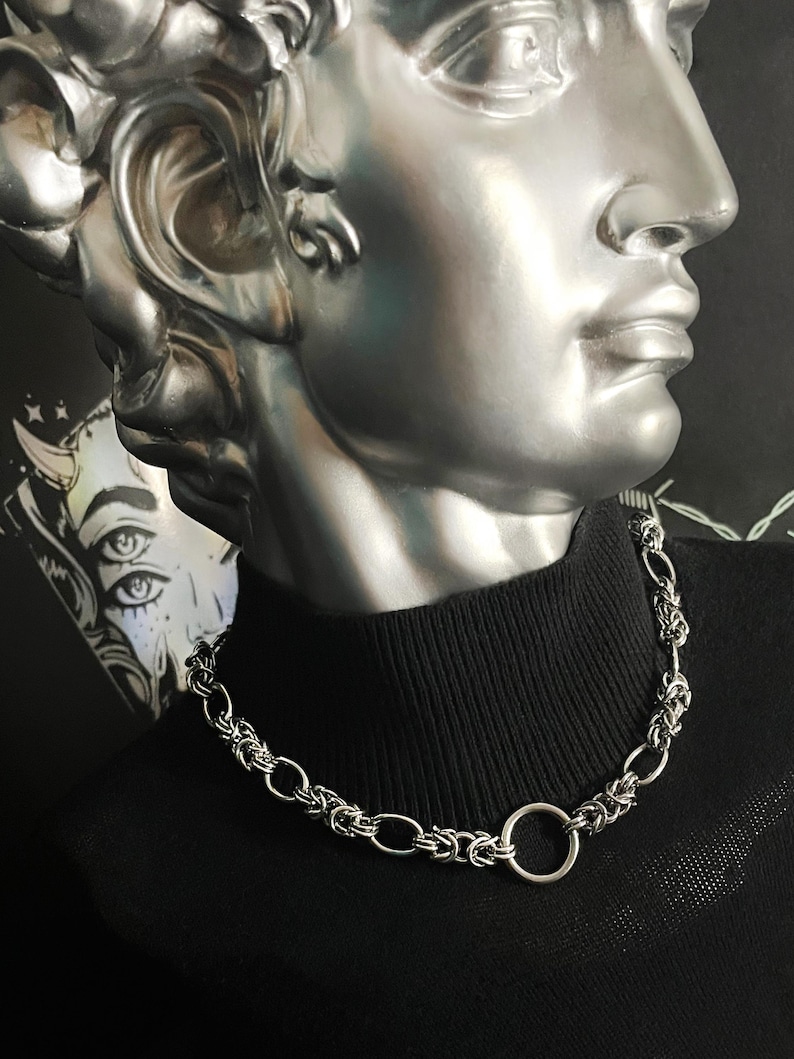 K&ouml;nnte beinhalten: Eine silberne Kettenkette mit einem gro&szlig;en, runden, silbernen Ringverschluss. Die Kette wird von einer Person getragen, die ein schwarzes Rollkragenhemd tr&auml;gt.