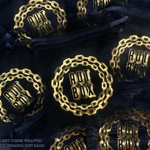 K&ouml;nnte beinhalten: Mehrere schwarze Organza-Geschenkbeutel mit einem goldfarbenen Kettenmuster aus Folie und dem Text "AUT BOX" in einem Kreis.