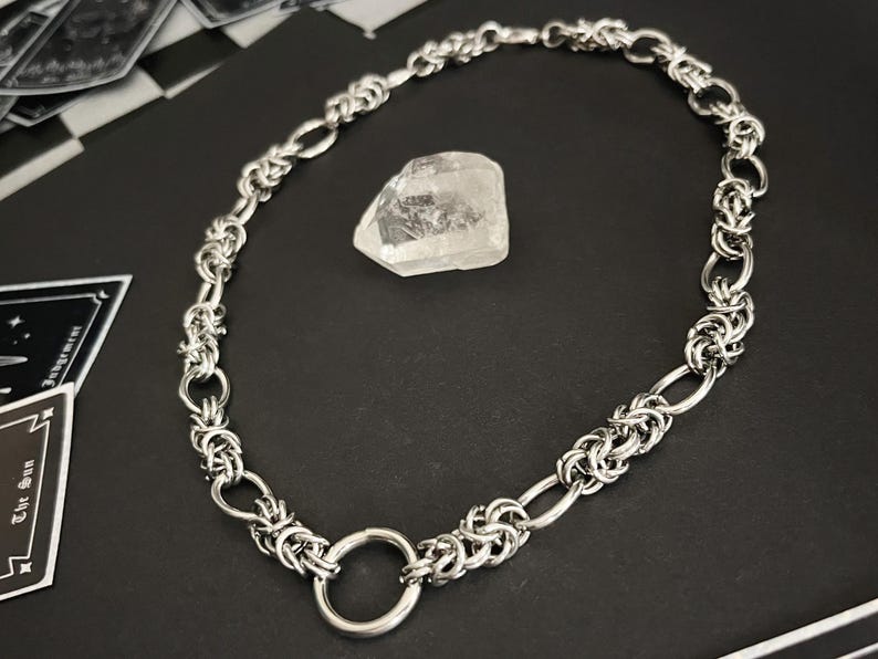 K&ouml;nnte beinhalten: Eine silberne Kettenkette mit einem gro&szlig;en, runden, offenen Ringverschluss. Die Kette besteht aus ineinandergreifenden Metallringen.