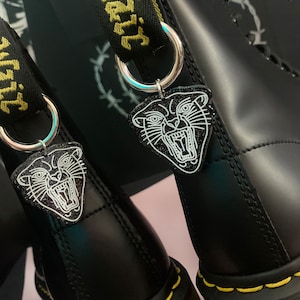 May include: Black leather boots with yellow stitching and a black panther charm attached to each boot. The charms are silver and have a white outline of a panther with an open mouth.