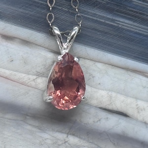 May include: A silver pendant necklace featuring a pear-shaped, pink gemstone. The gemstone is set in a silver bezel and hangs from a delicate silver chain. The background is a mix of white and blue tones.