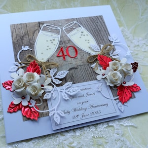 May include: A handmade 40th Ruby Wedding Anniversary card. The card features two champagne glasses, red '40' numerals, white paper flowers, red leaves, and a printed message. The card is decorated with white paper cutouts and twine bows.