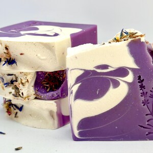 K&ouml;nnte beinhalten: Ein Stapel handgemachter Lavendelseifen mit einem Wirbelmuster in Violett- und Wei&szlig;t&ouml;nen. Die Seifen sind mit getrockneten Lavendelbl&uuml;ten verziert und haben ein Lavendelpflanzenmuster, das auf der Unterseite eingepr&auml;gt ist.